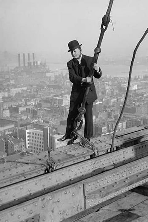 Cameraman in suit holds onto cable as he walks unharnessed over a skyscraper's steel girders